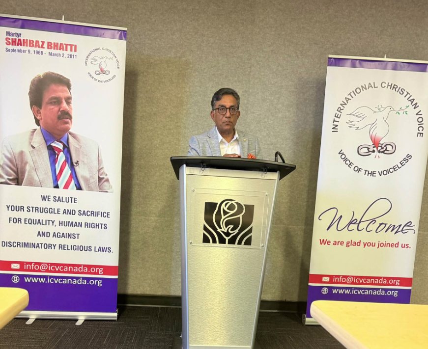 A speaker addresses the gathering between the Martyr Shahbaz Bhatti and International Christian Voice banners.